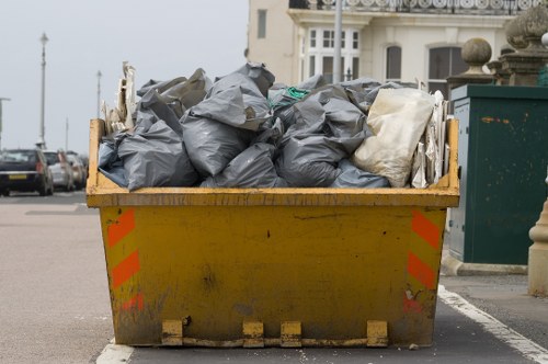 A large yellow skip bin filled with black and white rubbish bags, positioned on a paved street in an urban area. The bags appear to contain garden or household waste, and are stacked towards the top of the skip. The skip shows visible signs of dirt and wear, with orange reflective safety stickers on the sides. In the background, parked cars and residential buildings with white facades and large windows are partially visible, indicating a neighbourhood setting. The scene is captured during daylight hours, with overcast skies providing diffuse lighting. This image relates to rubbish removal services offered by Garden Clearance Peckham, serving the local area around SE15 postcode in Peckham.