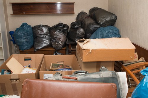 A collection of black and blue rubbish bags, some tightly tied and bulging, stacked on a wooden bench against a plain beige wall, with various cardboard boxes, some open revealing paper and packaging waste, placed in front of the bags. A large, partially flattened cardboard box rests on top of the boxes, and a metal trash bin or container is visible at the bottom right corner, all situated on an indoor surface that appears to be a garage or storage room. The scene depicts a typical rubbish clearance setup, with materials and waste awaiting removal or disposal, consistent with professional rubbish removal services in Peckham or the surrounding South London area.