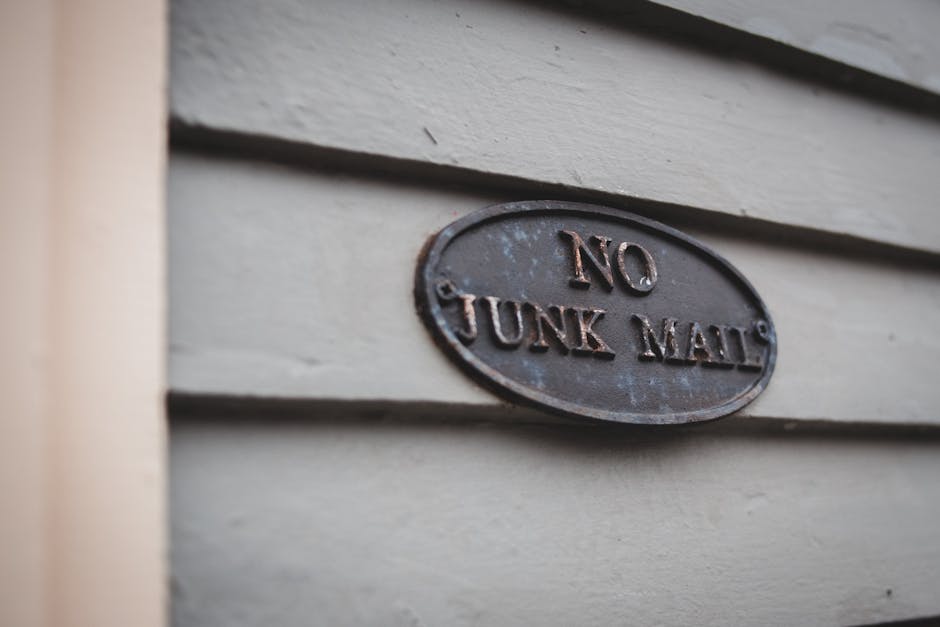A close-up photograph of an oval metal sign affixed to the exterior wall of a building. The sign is dark and weathered, with the raised embossed text reading 'NO JUNK MAIL' in capital letters. The wal