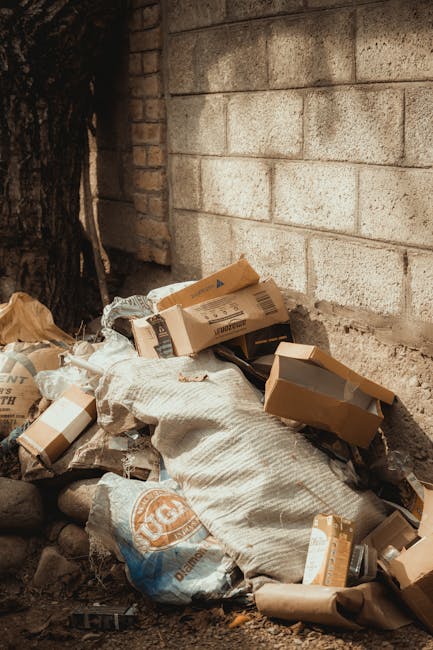A city street scene featuring a white garbage collection truck parked parallel to the pavement. The truck's rear compartment is open, revealing an empty, rusted interior with visible mechanical compon