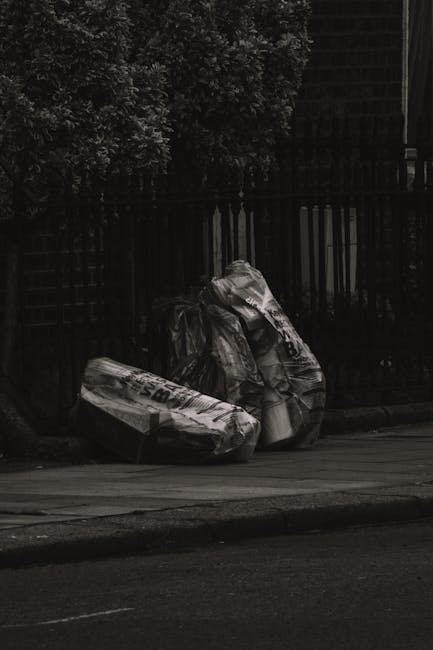 A collection of dumped household rubbish and waste materials is piled on a gravel surface in an outdoor area. The pile includes several black plastic trash bags partially torn open, revealing mixed wa