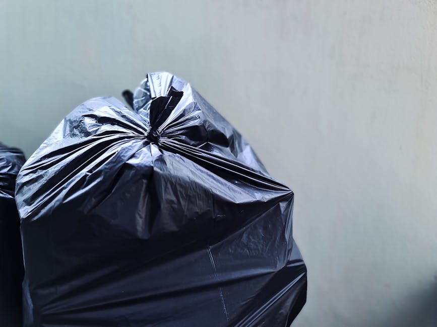 Close-up view of a tightly knotted black plastic rubbish bag with a glossy surface, placed in a pile of similar bags, against a neutral grey background. The bag's material appears crinkled and durable